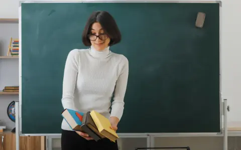 Nauczyciel początkujący z okularami trzyma stos książek przed zieloną tablicą.