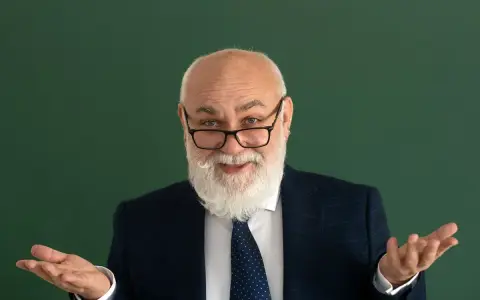 Uśmiechnięty starszy pan z brodą, w okularach i garniturze, gestykuluje, jakby wyjaśniał świadczenie kompensacyjne.
