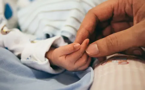 Dłoń rodzica obejmuje maleńką dłoń dziecka. To symbol miłości i troski, a także przygotowań do złożenia wniosku o urlop macierzyński.