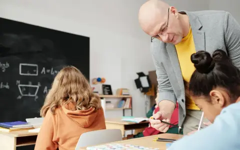 Nauczyciel pomaga uczniom podczas zajęć dydaktyczno wyrównawczych. Na tablicy wzory matematyczne.
