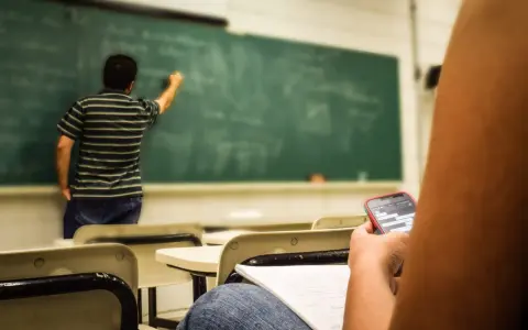 Nauczyciel pisze na tablicy, a uczeń w pierwszej ławce przegląda telefon. Obowiązki nauczyciela to nie tylko nauczanie, ale też pilnowanie porządku.