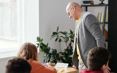 Nauczyciel pochyla się nad uczennicą, która pisze. W tle rośliny i regał z książkami. Czy to początek choroby zawodowej nauczycieli?