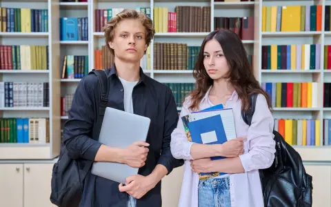 Dwóch studentów z książkami i laptopem na tle regałów z książkami.