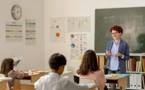 Uśmiechnięta dyrektor szkoły, w okularach, tłumaczy coś uczniom przy tablicy. W klasie wiszą kalendarze i mapy.