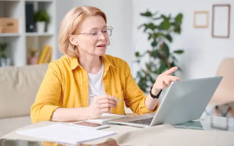 Kobieta w okularach i żółtej koszuli pracuje przy laptopie, trzymając długopis. To jej dodatek funkcyjny do pracy zdalnej.
