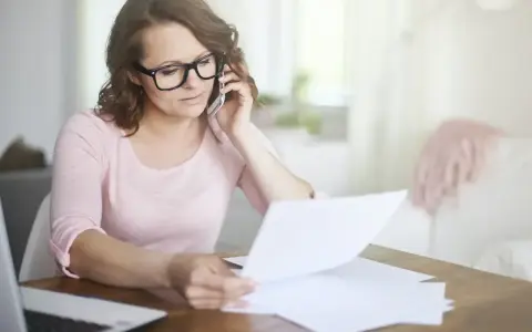 Kobieta w okularach przez telefon wyjaśnia sprostowanie świadectwa pracy, przeglądając dokumenty przy biurku.