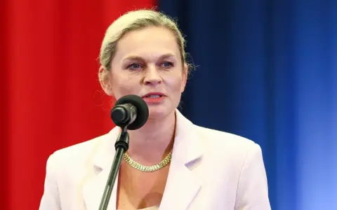 Kobieta w jasnej marynarce mówi do mikrofonu na tle czerwono-niebieskiego tła.