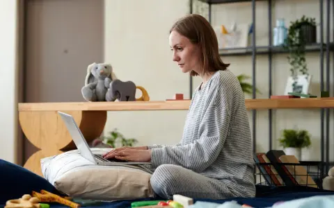 Kobieta na łóżku pisze wniosek o urlop rodzicielski na laptopie, otoczona zabawkami i pluszakami.