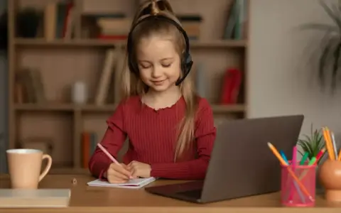 Uśmiechnięta dziewczynka w słuchawkach z mikrofonem pisze w zeszycie. To jej nauczanie indywidualne online, z laptopem przed sobą.