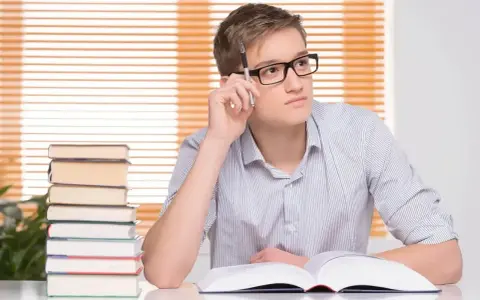 Młody mężczyzna w okularach, z długopisem w dłoni, zamyślony nad książką. Stos książek obok. Jego statut to student.