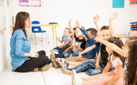 Nauczycielka siedzi na podłodze, a dzieci z podniesionymi rękami odpowiadają na pytania. Widać, że frekwencja w szkole jest wysoka, a uczniowie chętnie uczestniczą w lekcji.