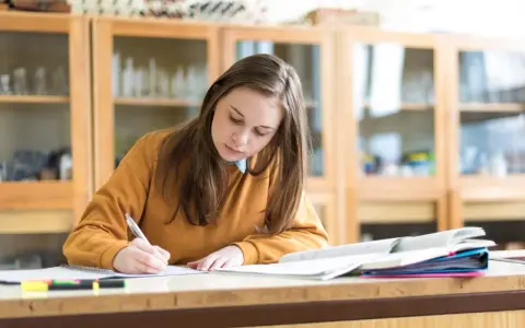 Dziewczyna pisze w zeszycie, przygotowując się do egzaminu. Marzy o stypendium szkolnym, które pomoże jej w dalszej nauce.