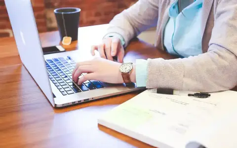 Osoba pisząca na laptopie, z kubkiem herbaty i notatkami na biurku.
