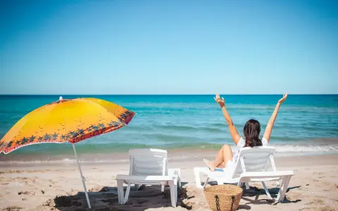 Kobieta cieszy się słońcem na plaży, w pełni wykorzystując swój urlop. Obok parasol i leżaki.