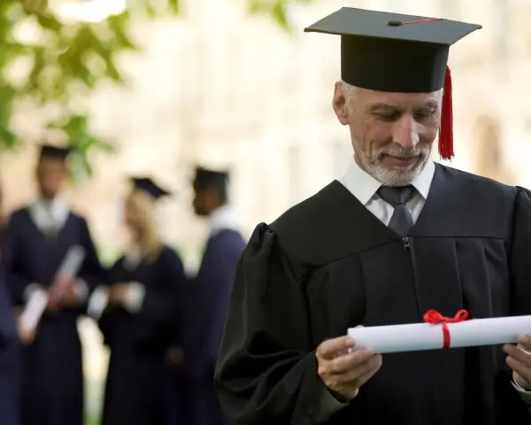 Starszy pan w todze z dyplomem zastanawia się, czy studia liczą się do stażu pracy. W tle inni absolwenci.