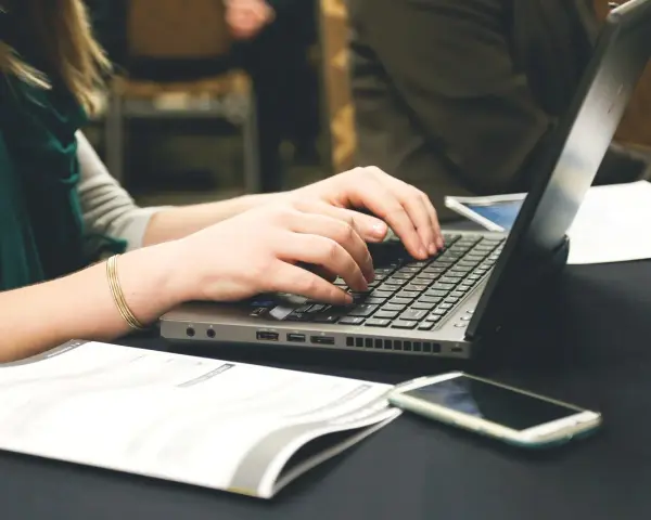 Dłonie piszą na laptopie, obok leży telefon i dokumenty. Może to być praca nad statutem szkoły.