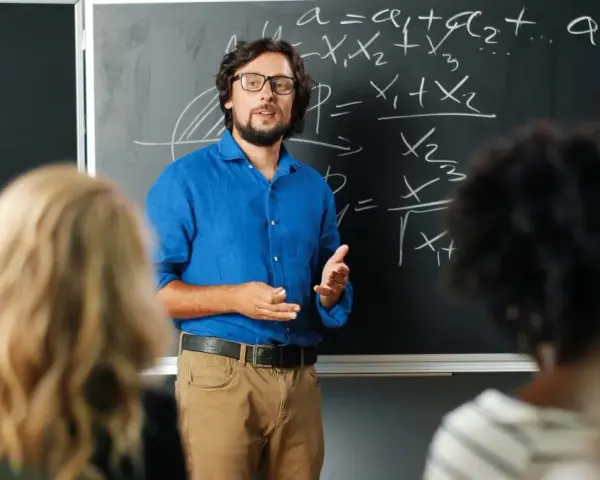 Nauczyciel w niebieskiej koszuli tłumaczy matematykę przy tablicy. Czy nauczyciel jest funkcjonariuszem publicznym? To pytanie nurtuje wielu.