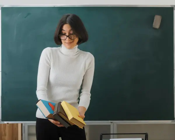 Nauczyciel początkujący z okularami trzyma stos książek przed zieloną tablicą.