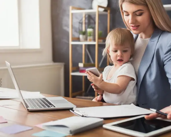 Praca na urlopie macierzyńskim? Młoda mama z dzieckiem przy biurku z laptopem i tabletem, pokazująca, że można łączyć obowiązki.