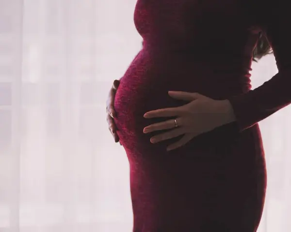 Kobieta w ciąży, z dłońmi na brzuchu, w ciemnoczerwonej sukience. Myśli o zasiłku macierzyńskim po ustaniu zatrudnienia.