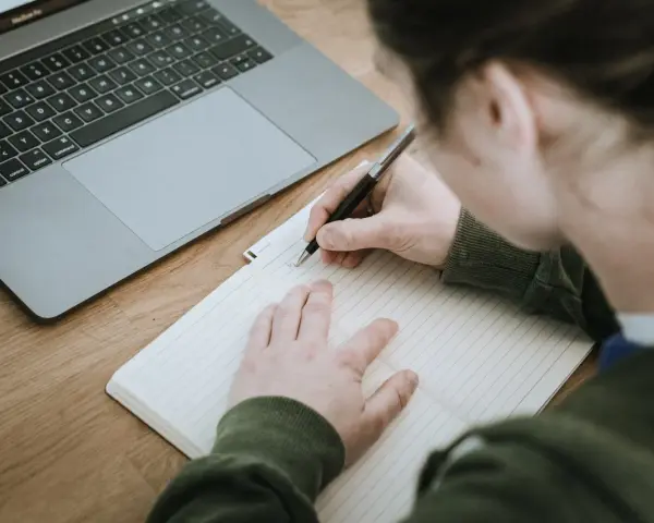 Osoba pisze w zeszycie, obok leży laptop. Powstaje opinia o uczniu, która zostanie zapisana.