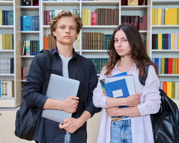Dwóch studentów z książkami i laptopem na tle regałów z książkami.