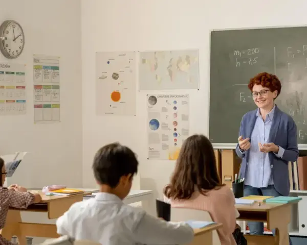 Uśmiechnięta dyrektor szkoły, w okularach, tłumaczy coś uczniom przy tablicy. W klasie wiszą kalendarze i mapy.