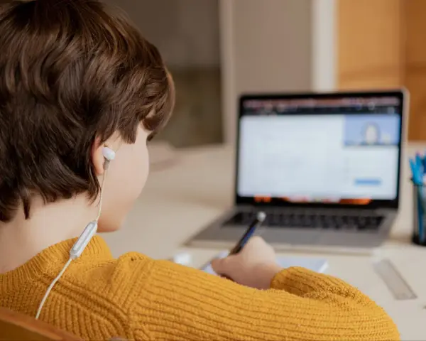 Chłopiec w żółtym swetrze uczy się zdalnie, pisząc notatki. Czy uczniowie dostaną laptopy, by móc tak pracować?