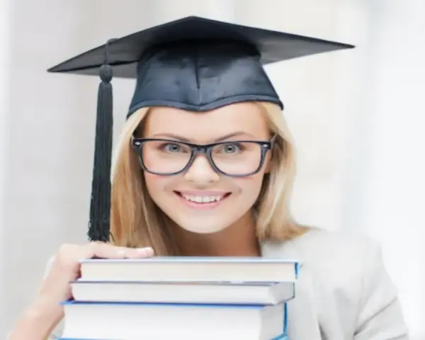 Uśmiechnięta studentka w birecie i okularach trzyma stos książek, zastanawiając się, ile trwają studia podyplomowe.