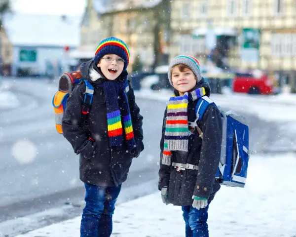 Dwóch chłopców w kolorowych czapkach i szalikach, gotowych na ferie 2027. Śnieg pada, a oni z plecakami czekają na przygodę.