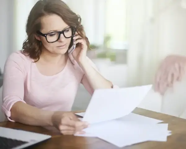 Kobieta w okularach przez telefon wyjaśnia sprostowanie świadectwa pracy, przeglądając dokumenty przy biurku.