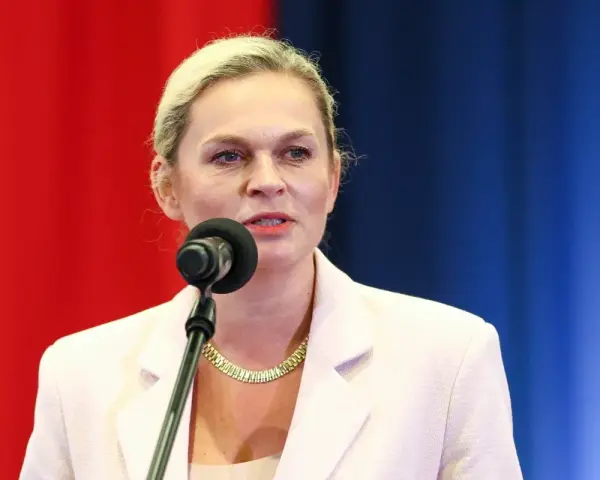 Kobieta w jasnej marynarce mówi do mikrofonu na tle czerwono-niebieskiego tła.