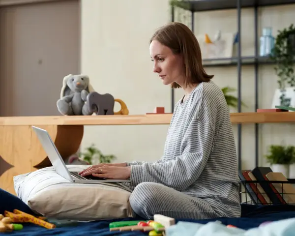 Kobieta na łóżku pisze wniosek o urlop rodzicielski na laptopie, otoczona zabawkami i pluszakami.