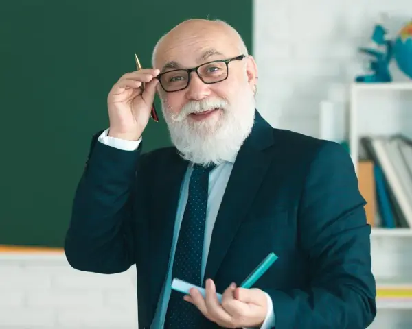 Uśmiechnięty starszy nauczyciel w okularach, z białą brodą, trzymający długopis. Myśli o zasłużonych emeryturach dla nauczycieli.