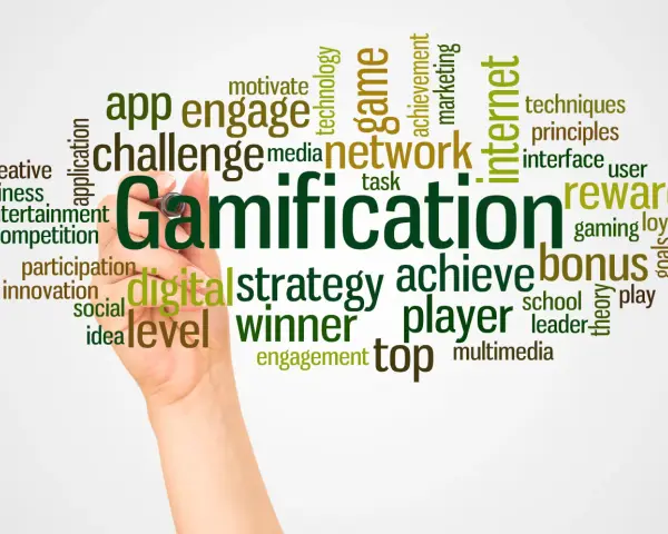 Ręka pisze słowo "Gamification" otoczone chmurą powiązanych terminów, takich jak "gra", "nagroda", "bonus", "strategia", "motywacja" i "sieć". Grywalizacja w akcji!