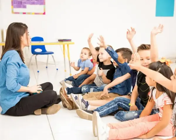 Nauczycielka siedzi na podłodze, a dzieci z podniesionymi rękami odpowiadają na pytania. Widać, że frekwencja w szkole jest wysoka, a uczniowie chętnie uczestniczą w lekcji.