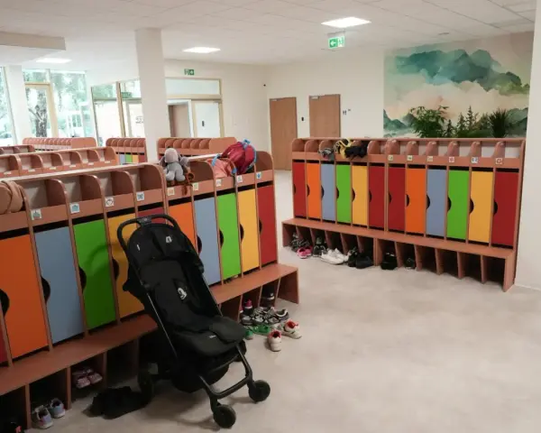 Kolorowe szafki i ławki w zespole szkolno-przedszkolnym, gotowe na przyjęcie dzieci. Wózek spacerowy czeka na swojego małego pasażera.