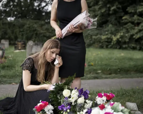 Smutna kobieta w czarnej sukience klęczy przy grobie, ocierając łzy. Obok stoi druga kobieta z bukietem kwiatów. To obraz żałoby, trudny urlop macierzyński po poronieniu.