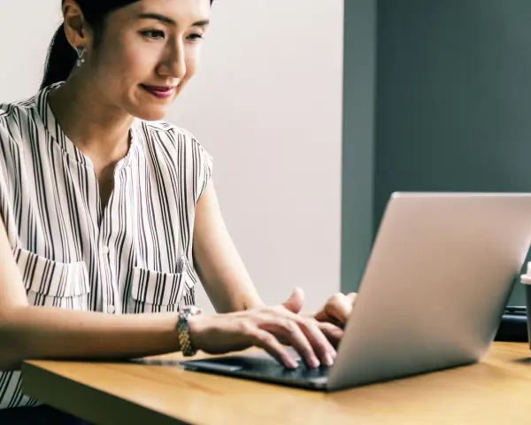 Kobieta uśmiecha się, pracując przy laptopie. Jej przygotowanie pedagogiczne online przebiega sprawnie.