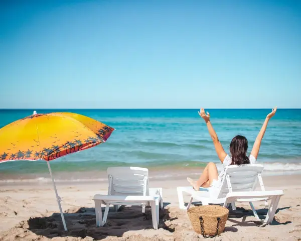 Kobieta cieszy się słońcem na plaży, w pełni wykorzystując swój urlop. Obok parasol i leżaki.
