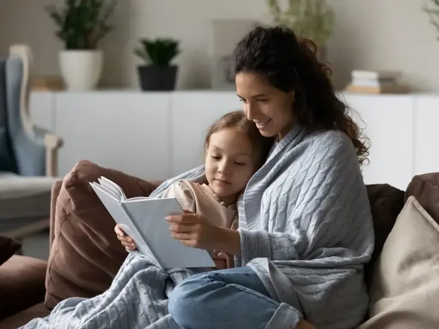 Ciepła opieka na dziecko 2 dni. Mama czyta bajkę córeczce, otulone kocem na kanapie.