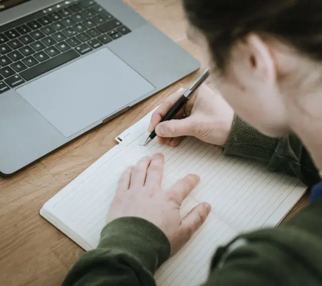 Osoba pisze w zeszycie, obok leży laptop. Powstaje opinia o uczniu, która zostanie zapisana.