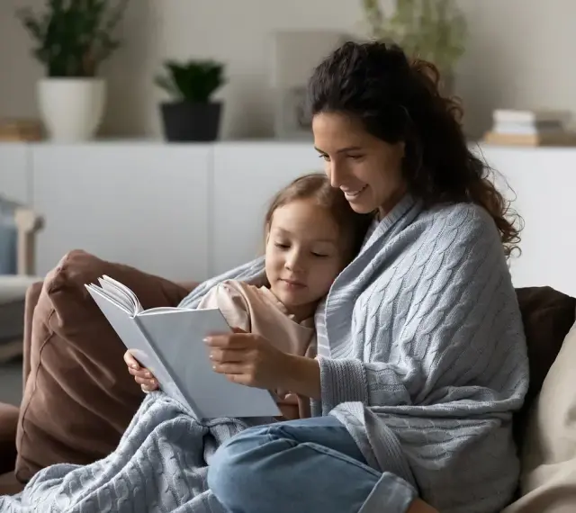 Ciepła opieka na dziecko 2 dni. Mama czyta bajkę córeczce, otulone kocem na kanapie.