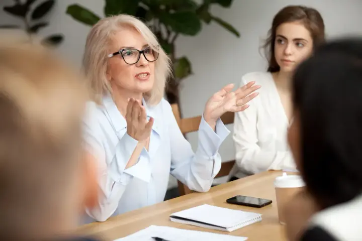Kobieta w okularach gestykuluje, prowadząc dyskusję podczas rady pedagogicznej.