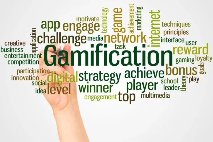 Ręka pisze słowo "Gamification" otoczone chmurą powiązanych terminów, takich jak "gra", "nagroda", "bonus", "strategia", "motywacja" i "sieć". Grywalizacja w akcji!