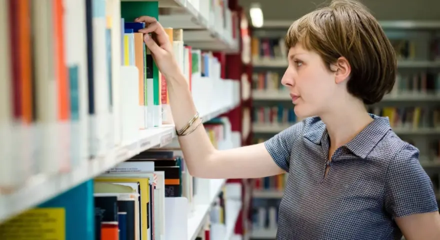 Kobieta, która wygląda jak bibliotekarz, wybiera książkę z półki w bibliotece.