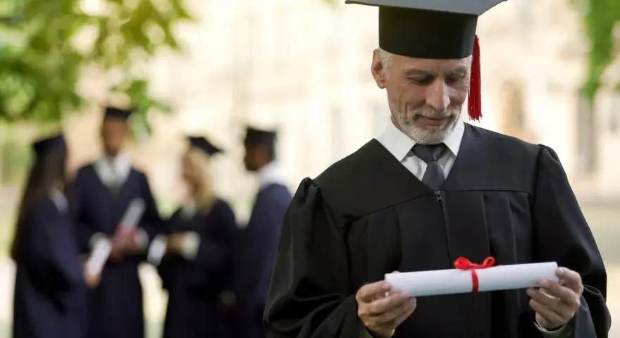 Starszy pan w todze z dyplomem zastanawia się, czy studia liczą się do stażu pracy. W tle inni absolwenci.