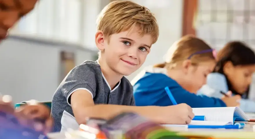 Uśmiechnięty chłopiec z blond włosami siedzi w klasie, otoczony nowymi przedmiotami w szkole. Obok niego inne dzieci piszą w zeszytach.