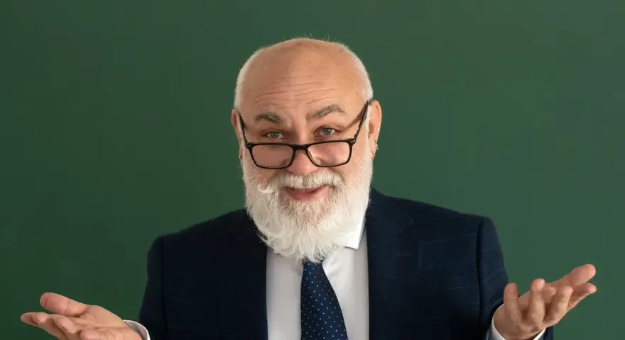 Uśmiechnięty starszy pan z brodą, w okularach i garniturze, gestykuluje, jakby wyjaśniał świadczenie kompensacyjne.