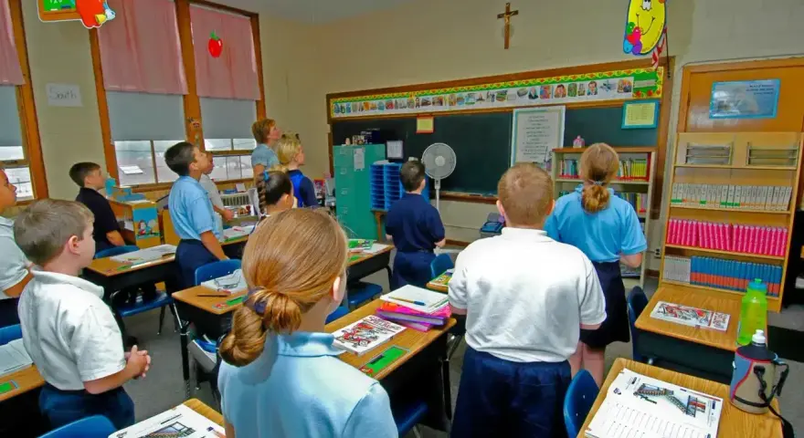 Dzieci słuchają katechety w klasie. Na ścianie wisi krzyż, a na tablicy kolorowe rysunki.